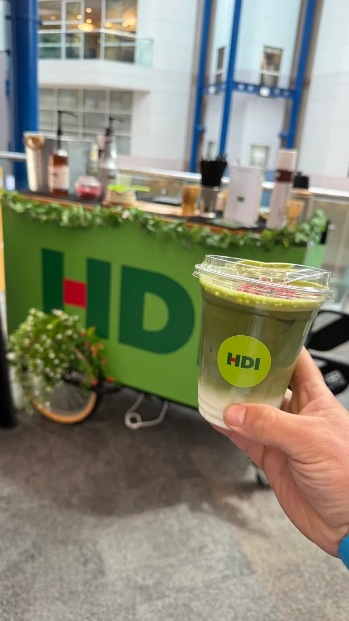 Hand holding a clear cup with a layered green iced drink and an HDI sticker, in front of an HDI-branded booth indoors.